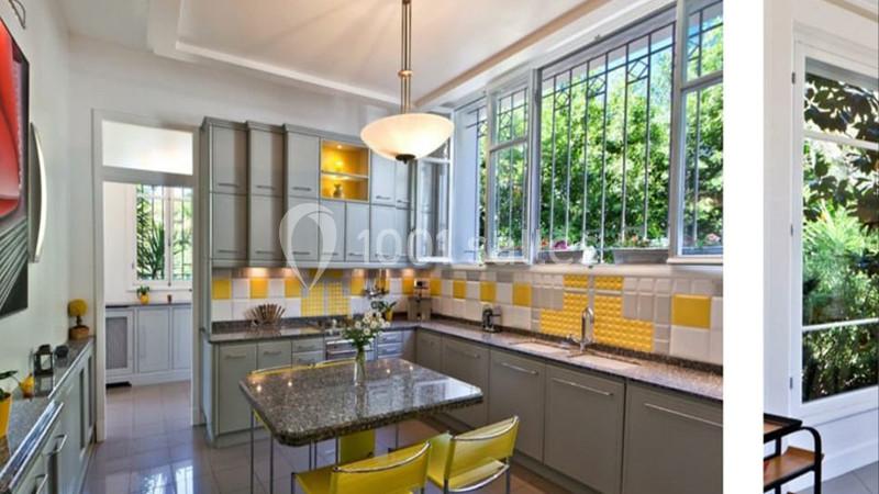 Cuisine lumineuse avec meubles gris, crédence jaune, îlot central en marbre et grandes fenêtres donnant sur un jardin.