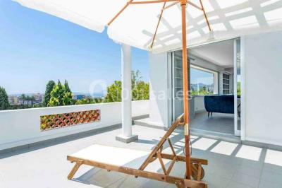 Vue panoramique sur une ville côtière avec des bâtiments, des arbres, la mer et des collines à l'horizon sous un ciel dégagé.