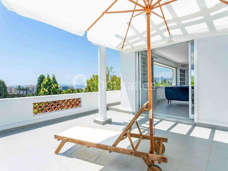 Transat en bois sous un parasol blanc sur une terrasse ensoleillée avec vue sur des arbres et une pièce ouverte.