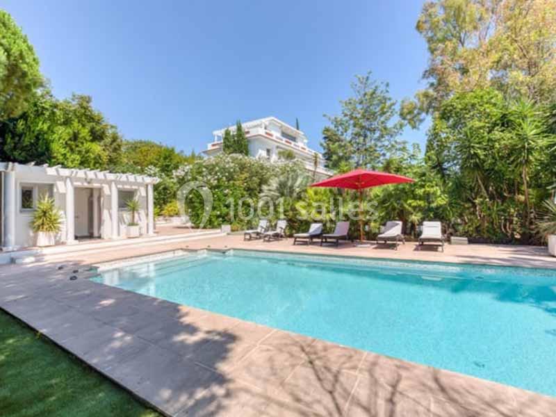 Piscine extérieure entourée de chaises longues, parasol rouge et végétation luxuriante, avec une maison blanche en arrière…
