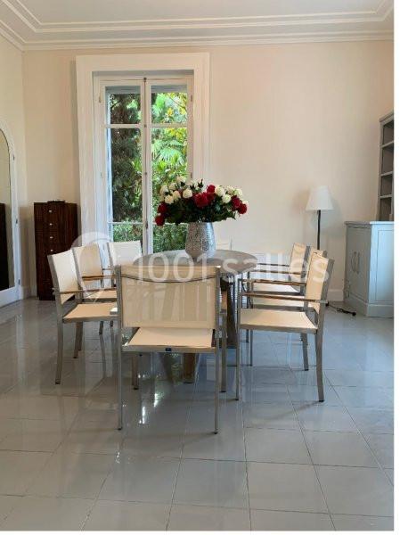 Salle à manger lumineuse avec table ronde, chaises blanches et vase de fleurs au centre, près d'une fenêtre donnant sur un…