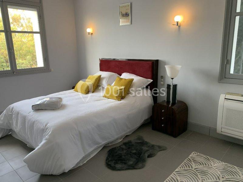 Chambre lumineuse avec lit double, coussins jaunes, murs clairs, deux fenêtres et éclairage mural.