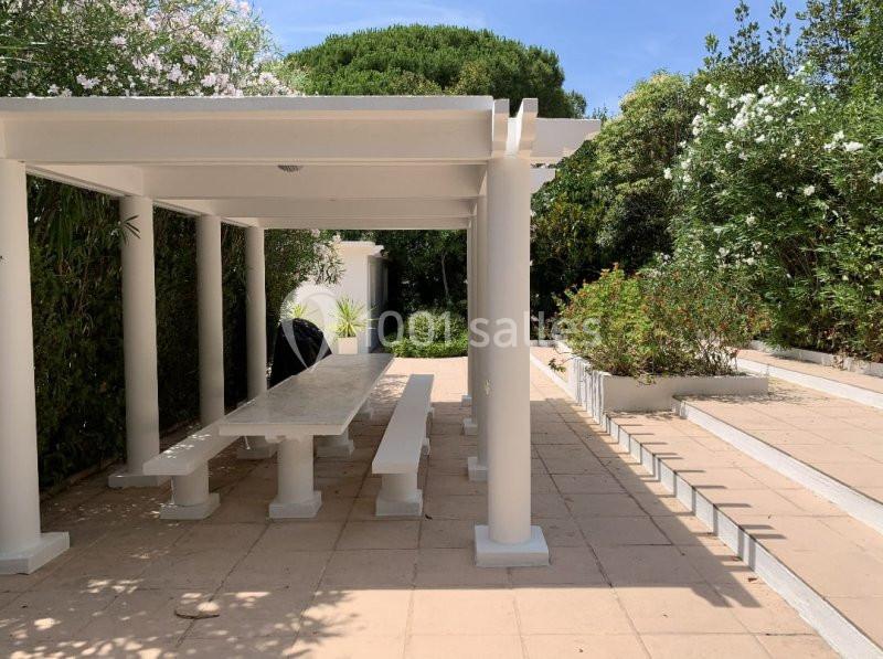 Pergola blanche avec table et bancs en pierre sur une terrasse entourée de végétation et de fleurs.