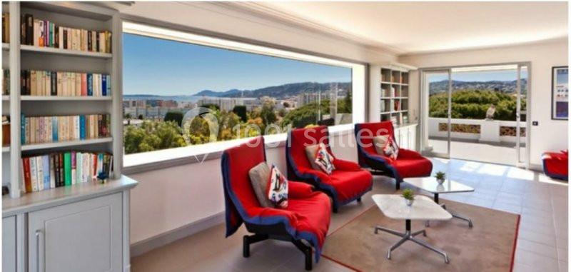 Salon lumineux avec grandes baies vitrées, trois fauteuils rouges, étagères remplies de livres et vue sur un paysage urbain.