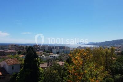 Vue panoramique sur une ville côtière avec des bâtiments, des arbres, la mer et des collines à l'horizon sous un ciel dégagé.