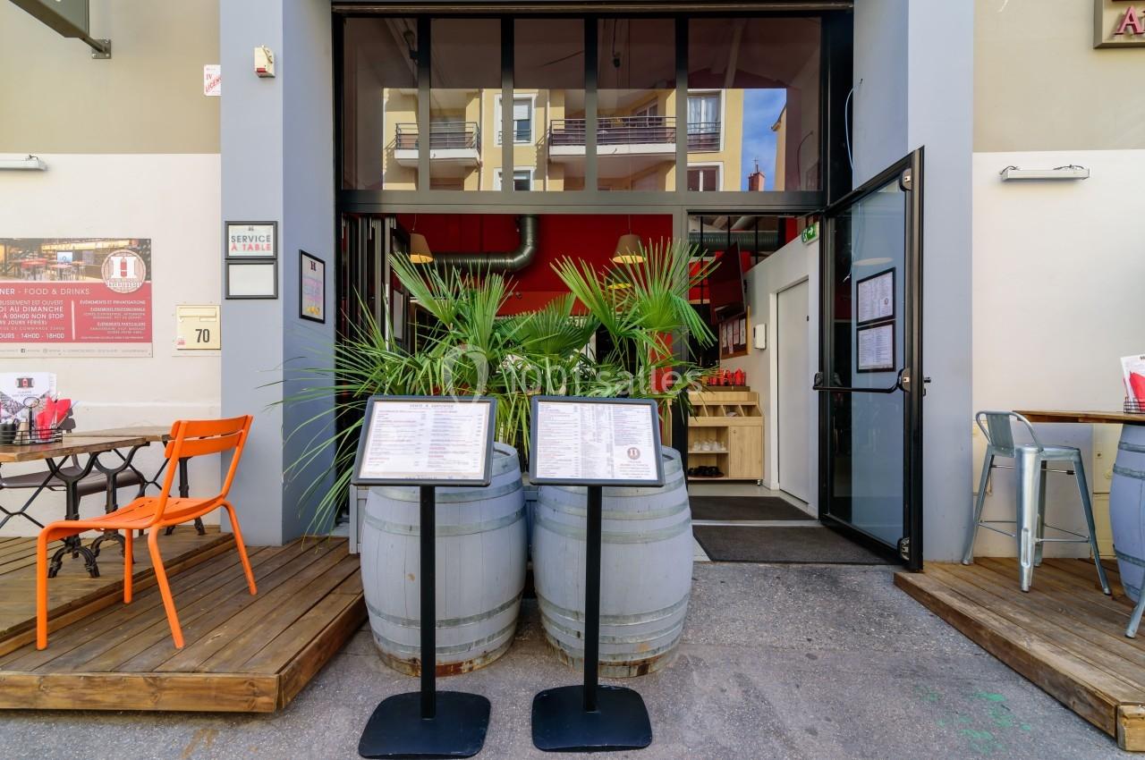 Entrée d'un restaurant avec menus affichés sur des tonneaux, terrasse aménagée et plantes décoratives.