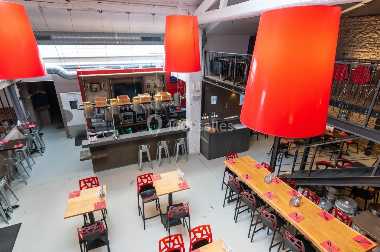 Salle de restaurant moderne avec tables en bois, chaises rouges, bar central et grandes suspensions rouges.
