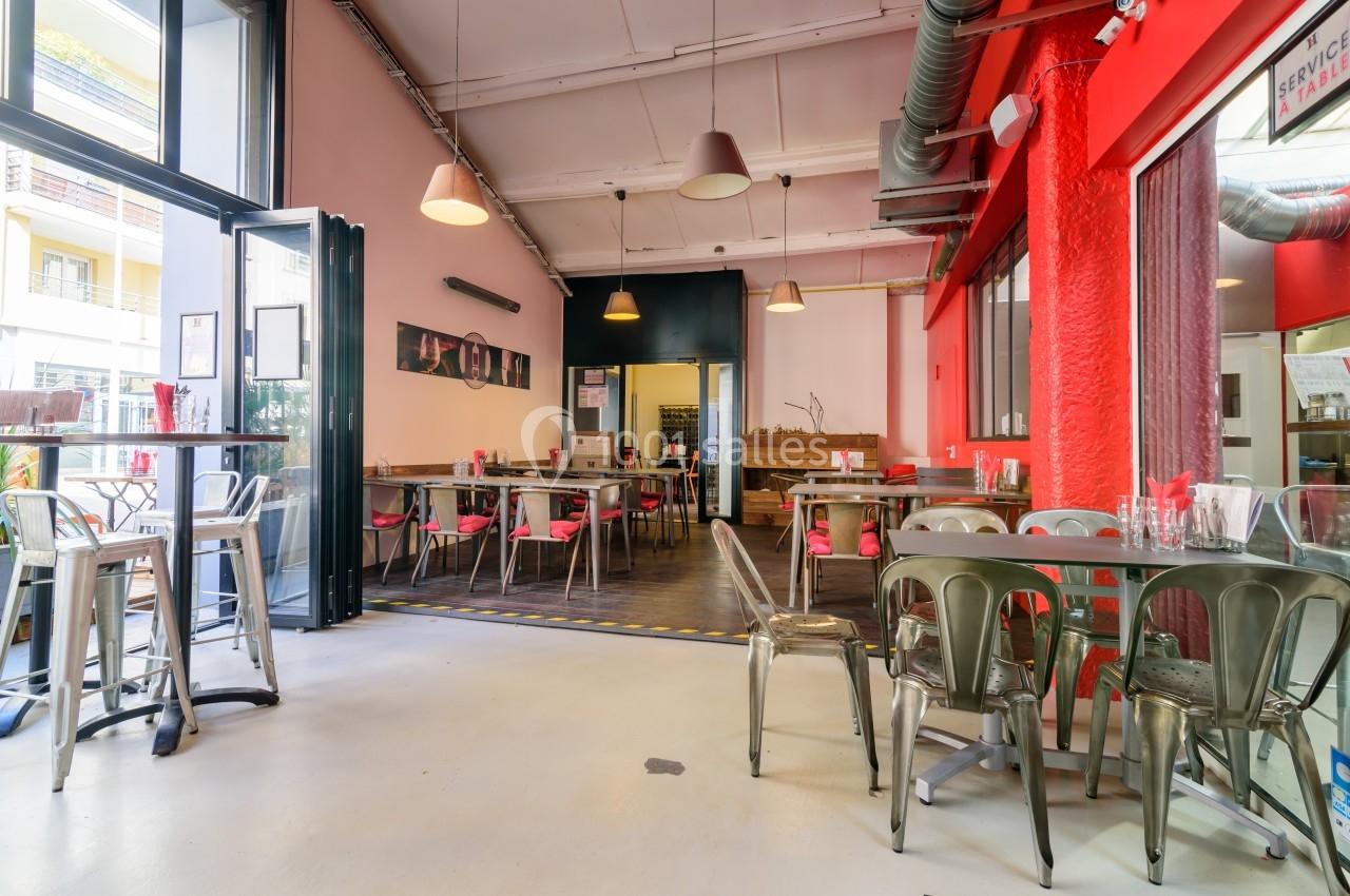 Salle de restaurant lumineuse avec des tables et chaises métalliques, murs rouges et roses, et grandes baies vitrées…