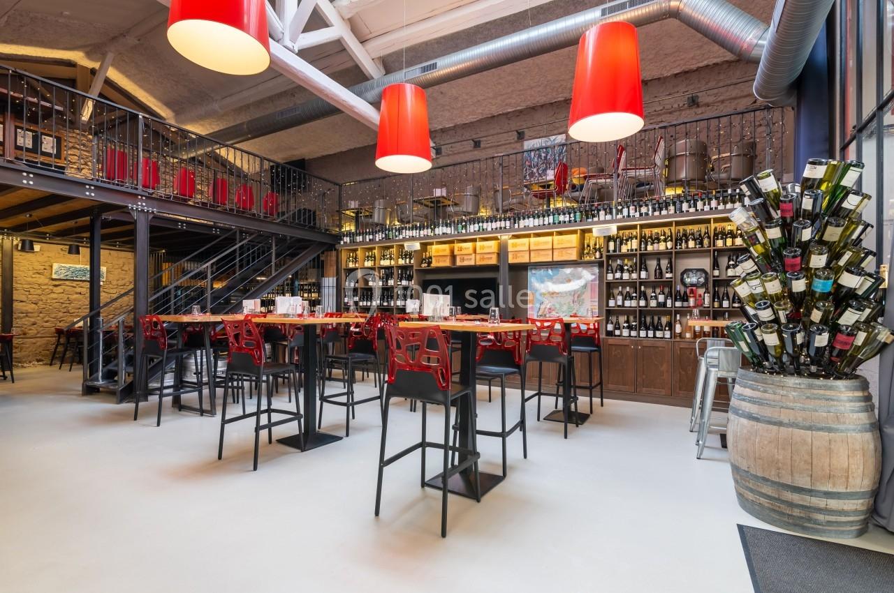 Intérieur d'un bar moderne avec tables hautes, étagères remplies de bouteilles de vin et lampes rouges suspendues.