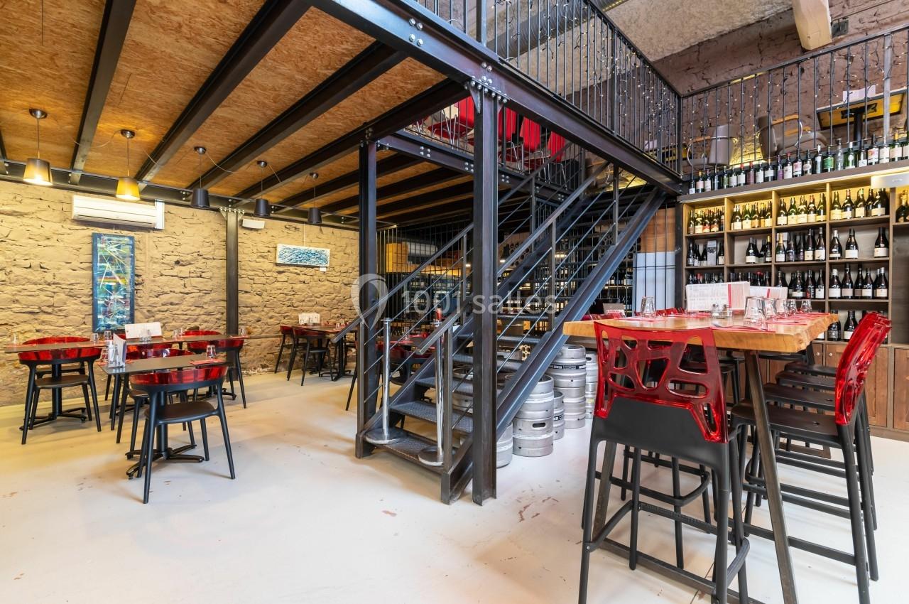 Salle de restaurant moderne avec tables, chaises rouges, escalier métallique et étagères remplies de bouteilles.