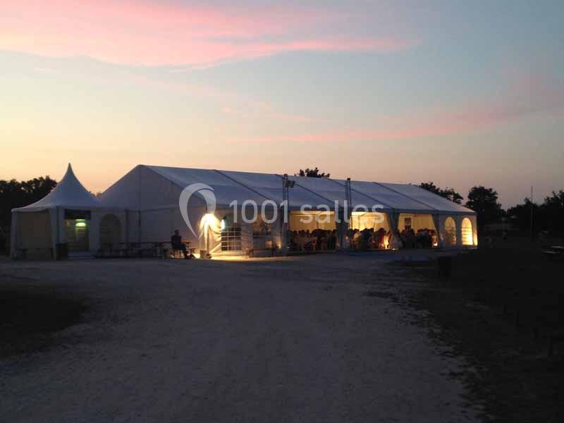Grande tente blanche éclairée de l'intérieur au crépuscule, installée sur un terrain en extérieur.