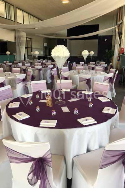 Salle de réception décorée avec des tables rondes dressées, nappes blanches, chemins de table violets et chaises assorties.
