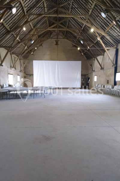 Salle spacieuse avec charpente en bois, tables et chaises disposées sur les côtés, écran blanc au fond.