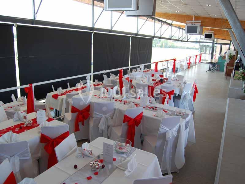 Salle décorée pour un événement avec des tables dressées, nappes blanches et chaises ornées de nœuds rouges.