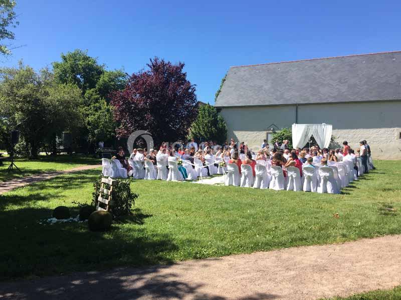 Invités assis en plein air lors d'une cérémonie, devant une arche décorée, dans un jardin par une journée ensoleillée.