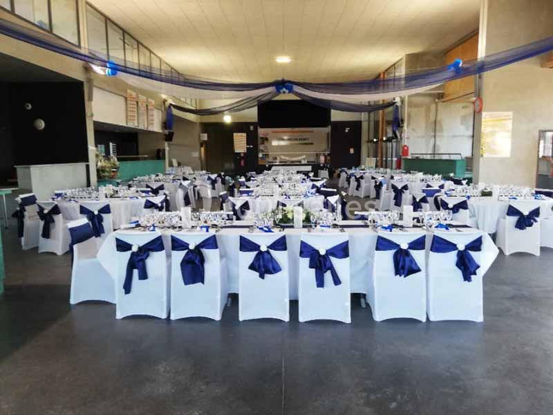 Salle de réception décorée avec des tables alignées, nappes blanches et chaises ornées de nœuds bleus.