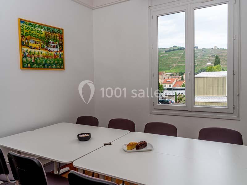 Salle de réunion lumineuse avec table blanche, chaises violettes, tableau coloré au mur et vue sur des vignobles par la…