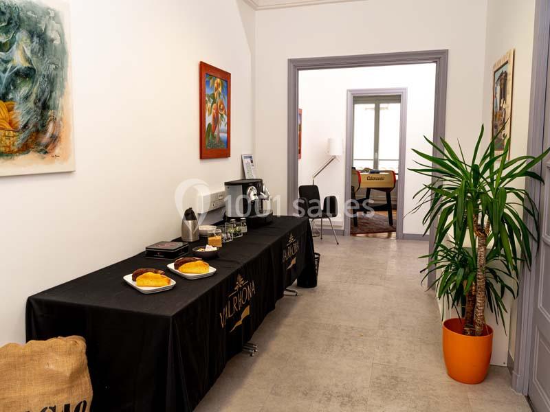Table avec viennoiseries, machine à café et boissons dans un couloir lumineux décoré de plantes et tableaux.