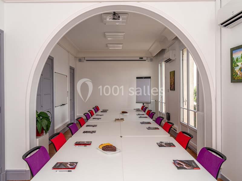 Salle de réunion lumineuse avec grande table blanche, chaises colorées, tableau blanc et documents posés.