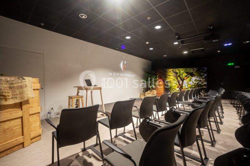 Salle de conférence avec chaises alignées, un pupitre en bois, un écran et une décoration murale colorée.