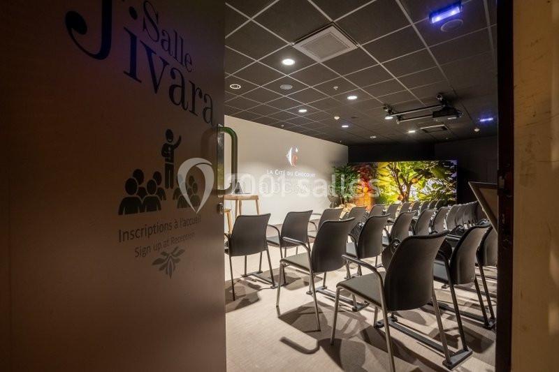 Salle de conférence avec rangées de chaises vides, écran et décor lumineux, vue depuis une porte entrouverte.