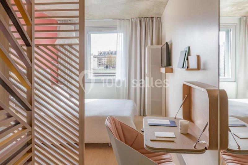 Chambre lumineuse avec lit, bureau en bois, chaise rose, cloison ajourée et grande fenêtre donnant sur l'extérieur.