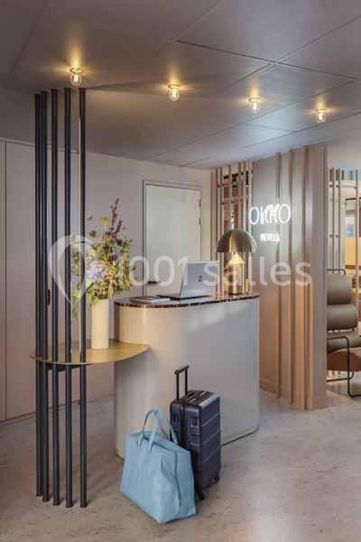 Accueil d'un hôtel moderne avec comptoir, lampe design, valises posées au sol et décoration florale.