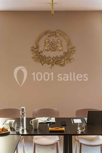 Salle de réunion avec table sombre, chaises roses, documents, tasses, et un emblème doré sur un mur beige.
