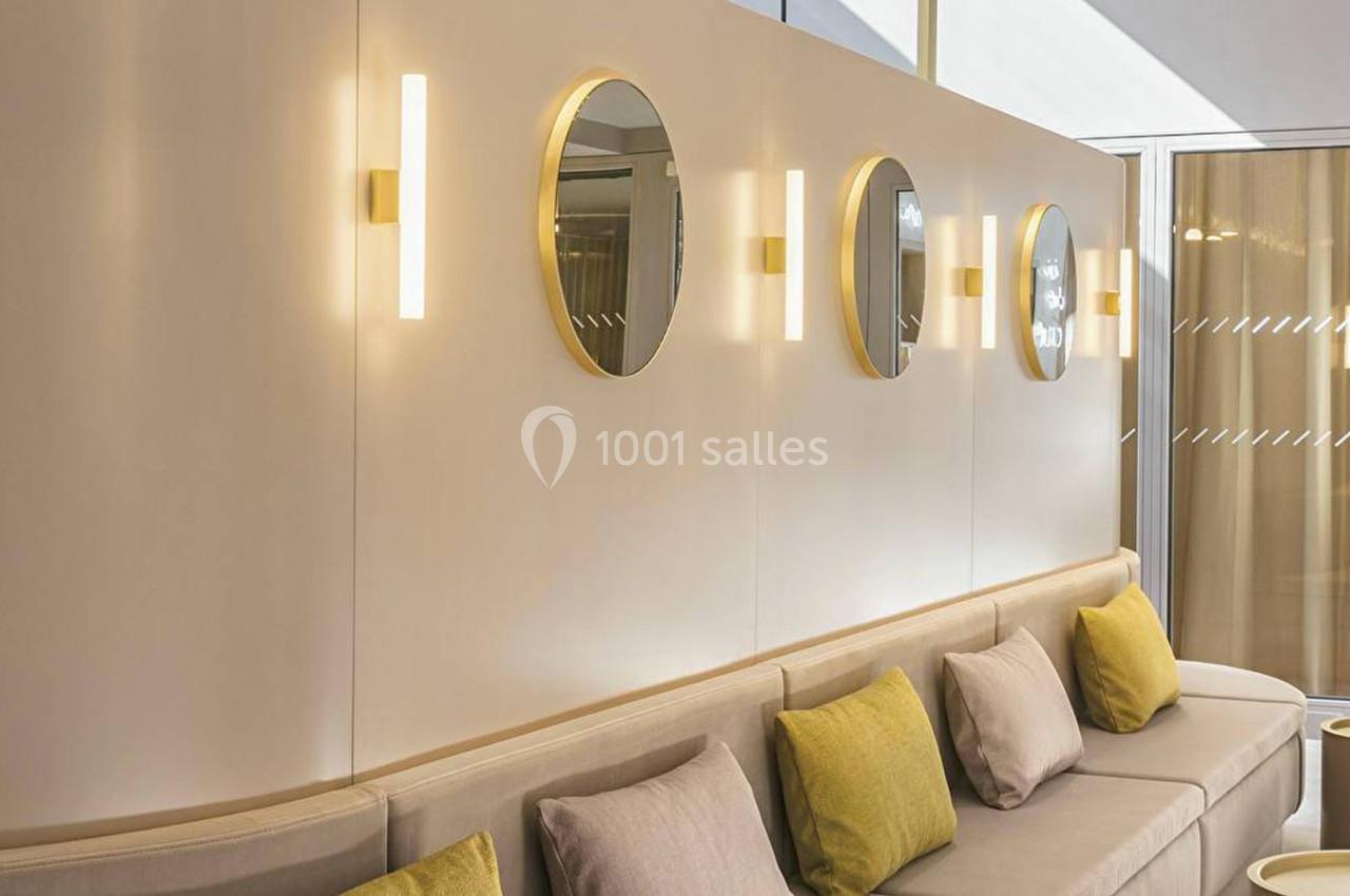 Mur beige avec miroirs ronds encadrés de doré, appliques lumineuses et canapé beige orné de coussins colorés.