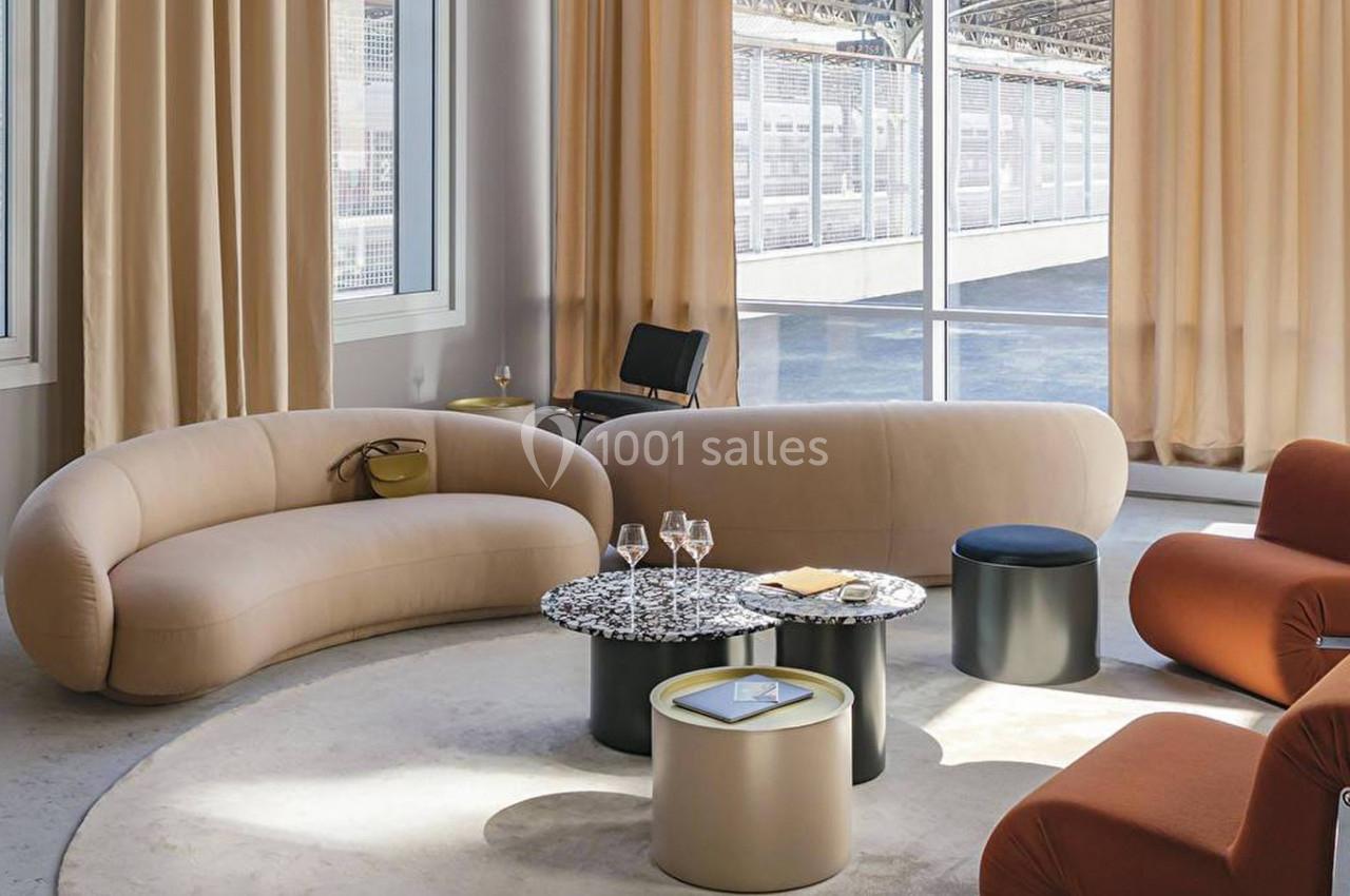 Salon moderne avec canapés beiges, fauteuils orange, tables basses rondes et grandes fenêtres laissant entrer la lumière.