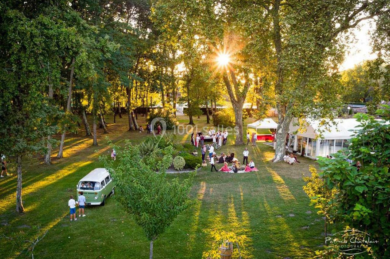 Vue aérienne d'un jardin ensoleillé avec des arbres, des invités rassemblés et un van vintage stationné.