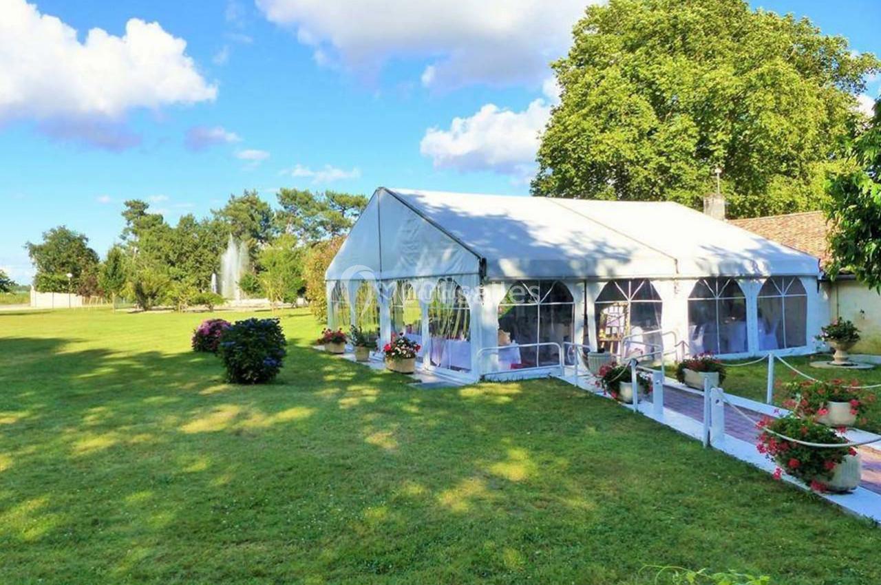 Tente blanche installée dans un jardin verdoyant avec des fleurs et des arbres sous un ciel partiellement nuageux.