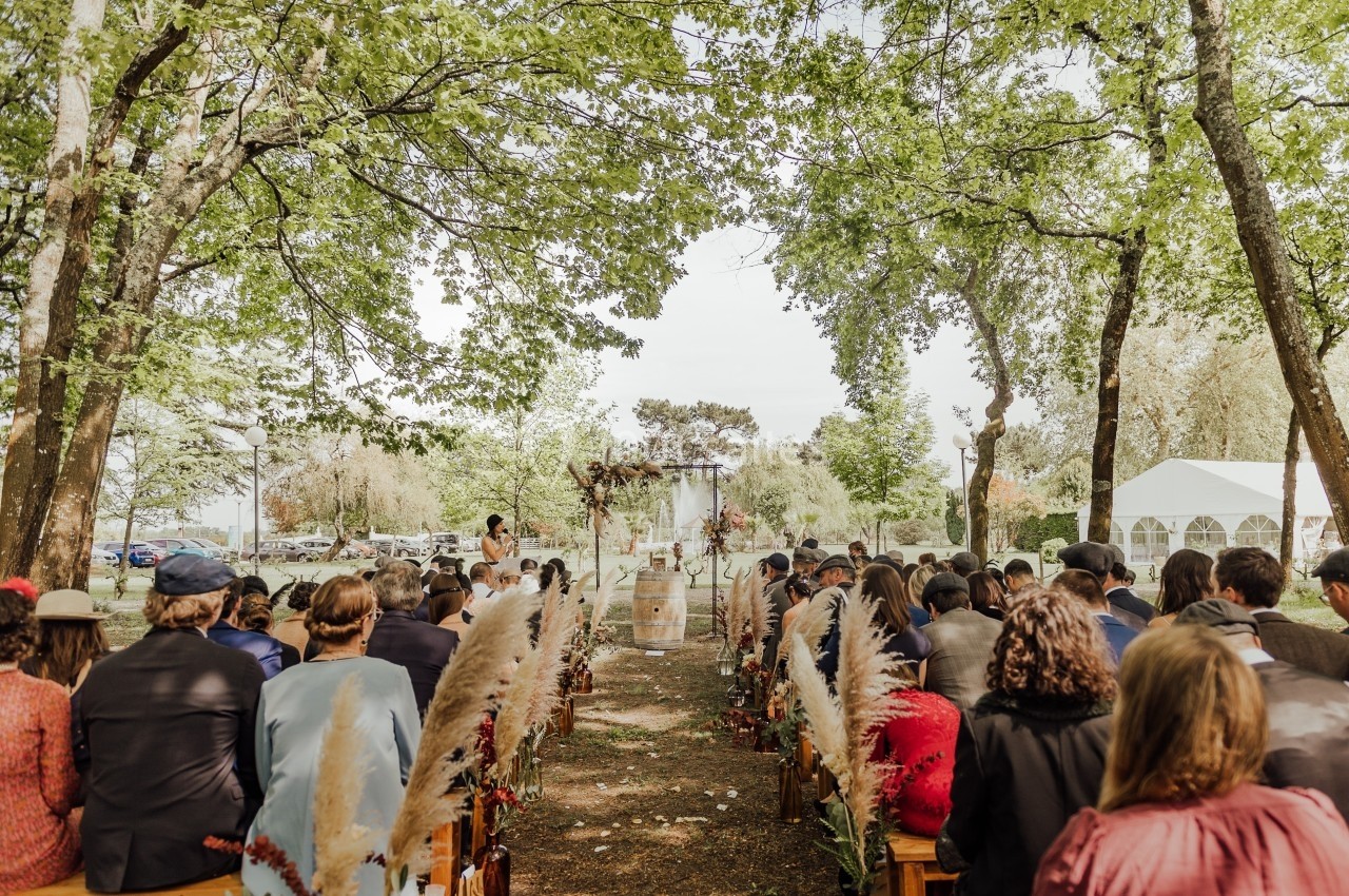 Cérémonie de mariage en plein air sous des arbres, avec des invités assis et une allée décorée de fleurs séchées.