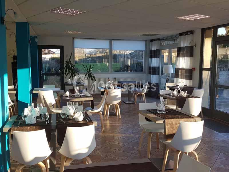 Salle de restaurant lumineuse avec tables dressées, chaises blanches et grandes baies vitrées donnant sur un extérieur…