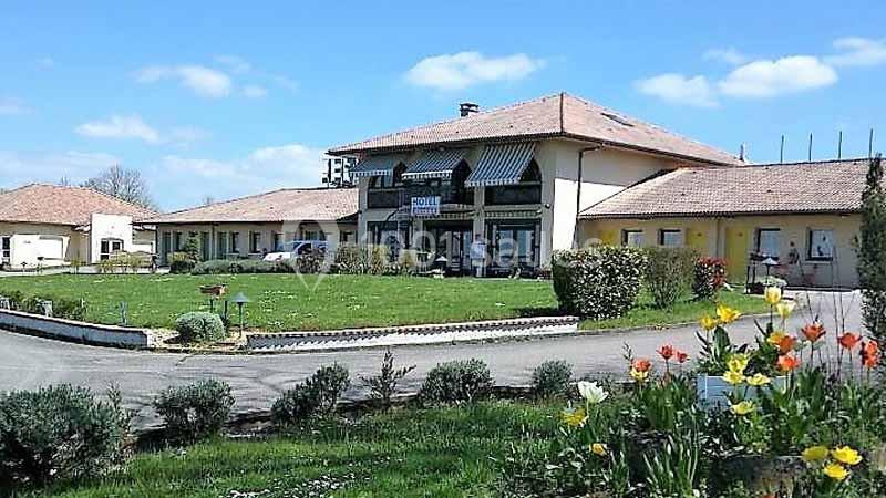 Bâtiment à un étage avec toit en tuiles, entouré d'un jardin fleuri et d'une allée pavée sous un ciel bleu.