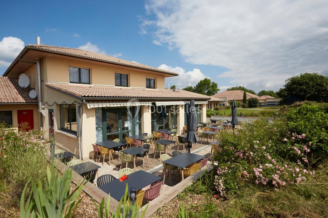 Terrasse extérieure d'un restaurant avec des tables et chaises, entourée de végétation et sous un ciel partiellement nuageux.