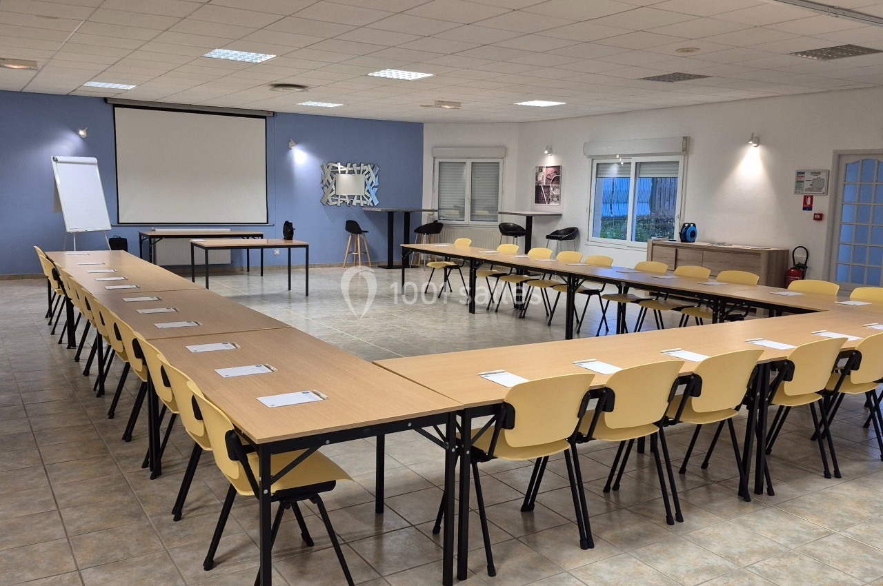 Salle de réunion lumineuse avec des tables disposées en U, des chaises jaunes et un écran de projection.