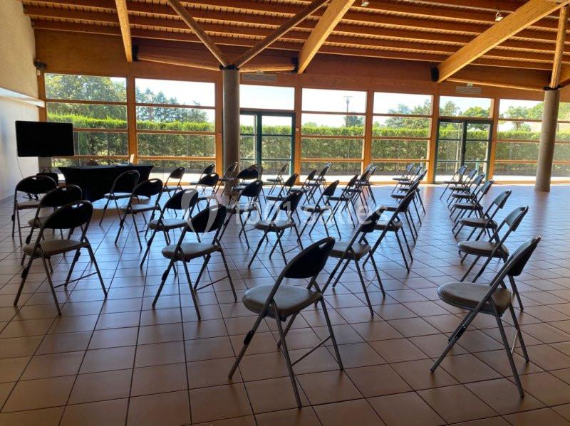 Salle lumineuse avec des rangées de chaises pliantes disposées face à un écran, grandes baies vitrées en arrière-plan.