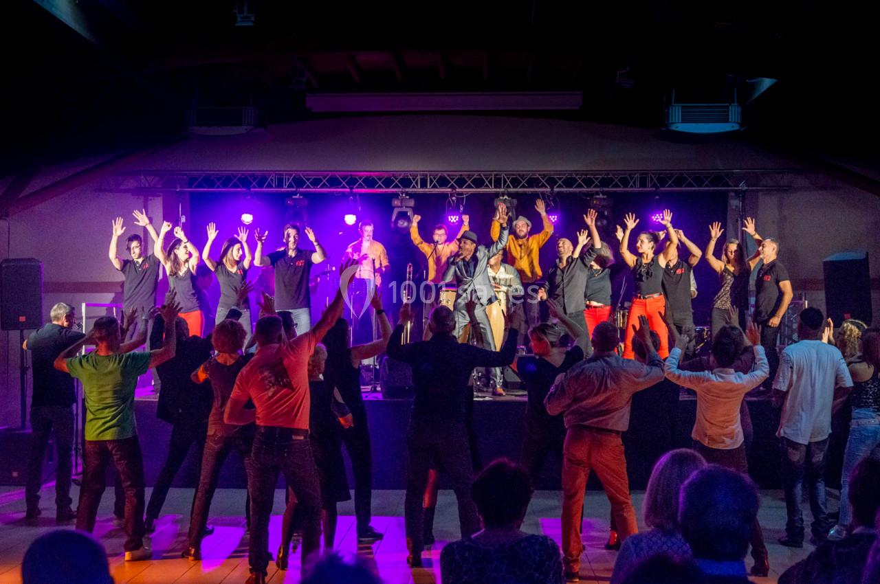 Groupe de musiciens et chanteurs sur scène avec un public debout levant les bras dans une ambiance festive en intérieur.