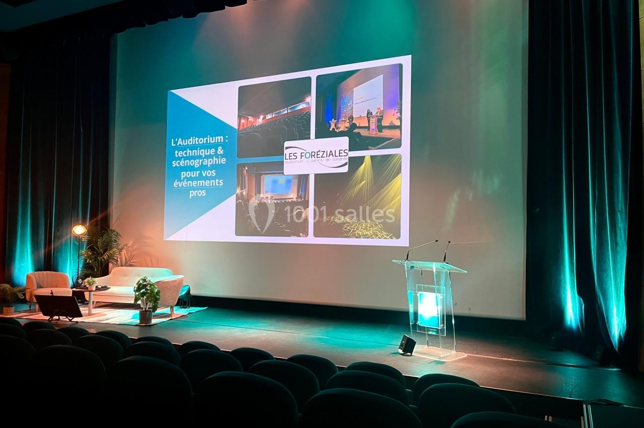 Scène d'auditorium vide avec écran de projection, fauteuils, pupitre et éclairage coloré.