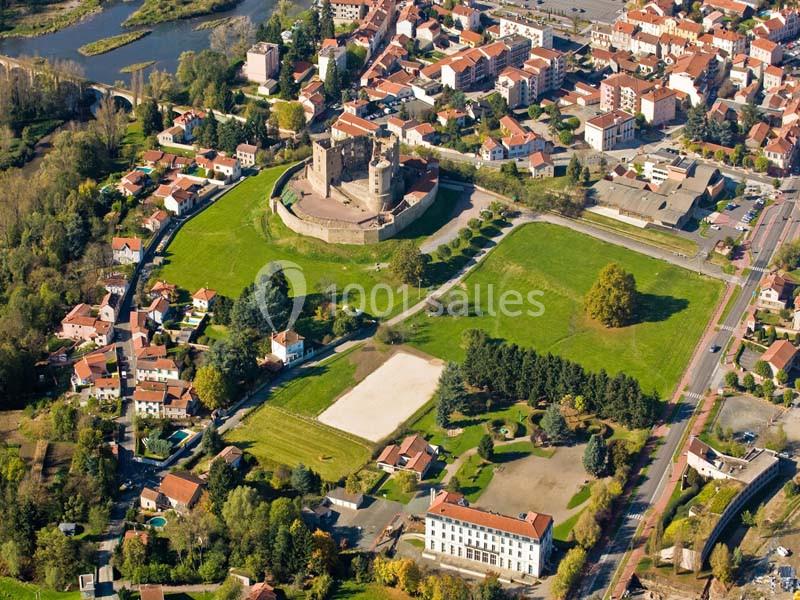 Vue aérienne d'un château médiéval entouré de verdure, au cœur d'une petite ville avec des habitations et des routes.