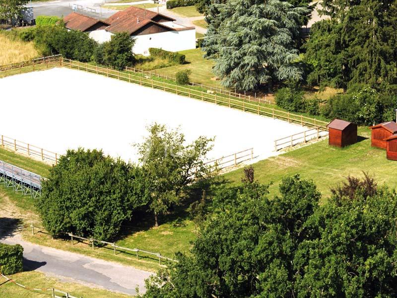 Vue aérienne d'une carrière équestre entourée de verdure, avec des arbres, des bâtiments et des abris en bois à proximité.