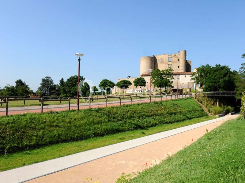 Vue d'un château médiéval en pierre entouré d'arbres et de chemins dans un paysage verdoyant sous un ciel bleu.