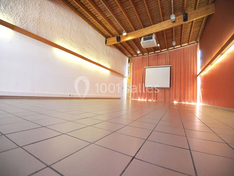 Salle vide avec sol carrelé, murs blancs et rouges, plafond en bois et écran de projection devant des rideaux.