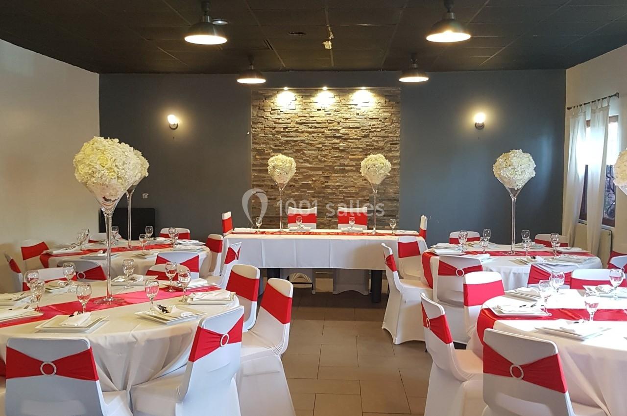 Salle de réception décorée avec des tables rondes, nappes blanches, chaises ornées de rubans rouges et centres de table…