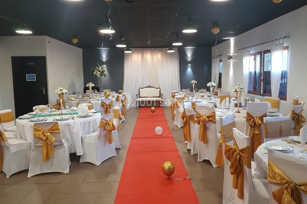 Salle de réception décorée avec des tables dressées, des chaises ornées de nœuds dorés et un tapis rouge central.