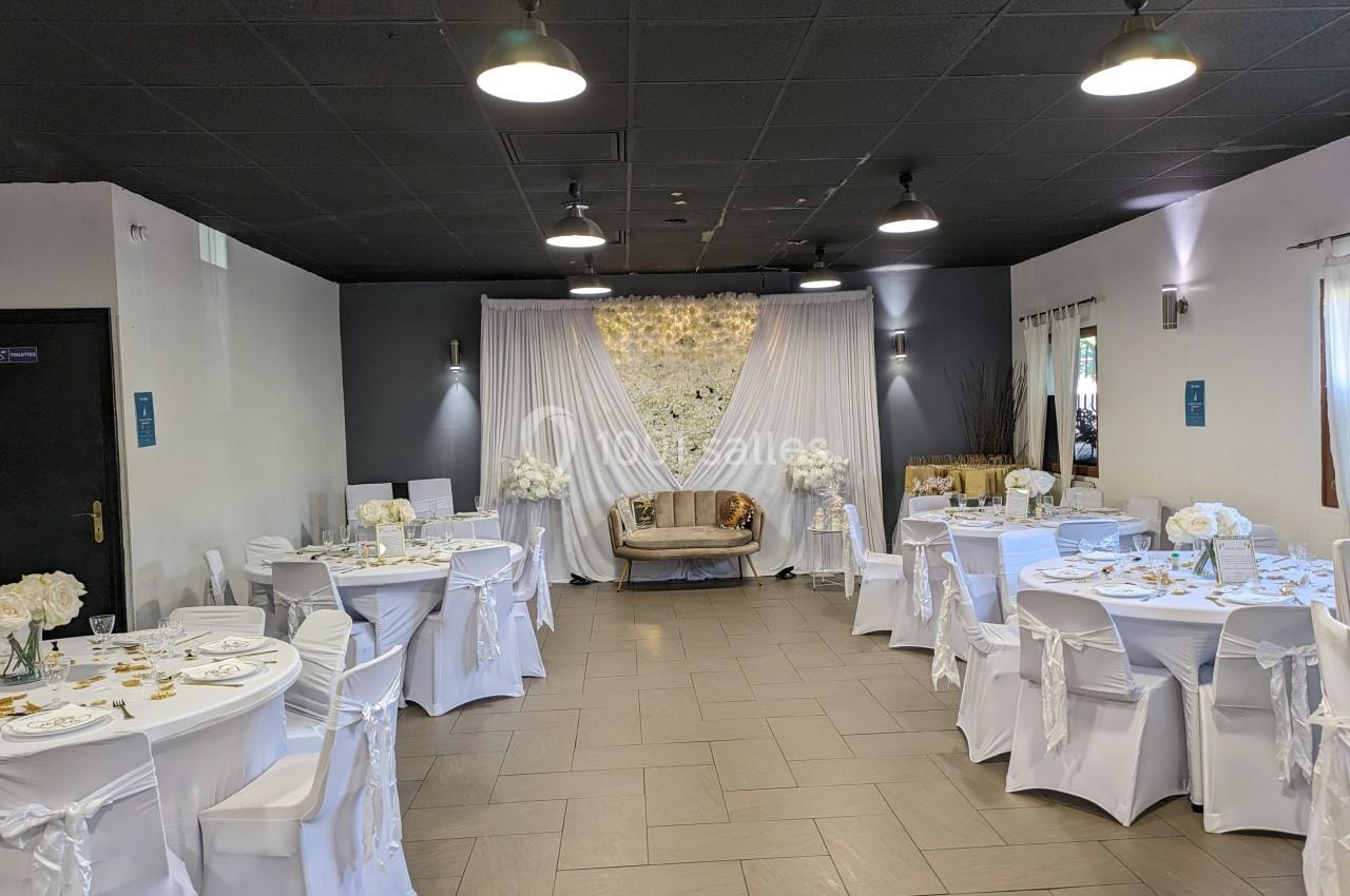 Salle de réception décorée en blanc avec des tables rondes, des chaises habillées et un canapé devant un mur fleuri.