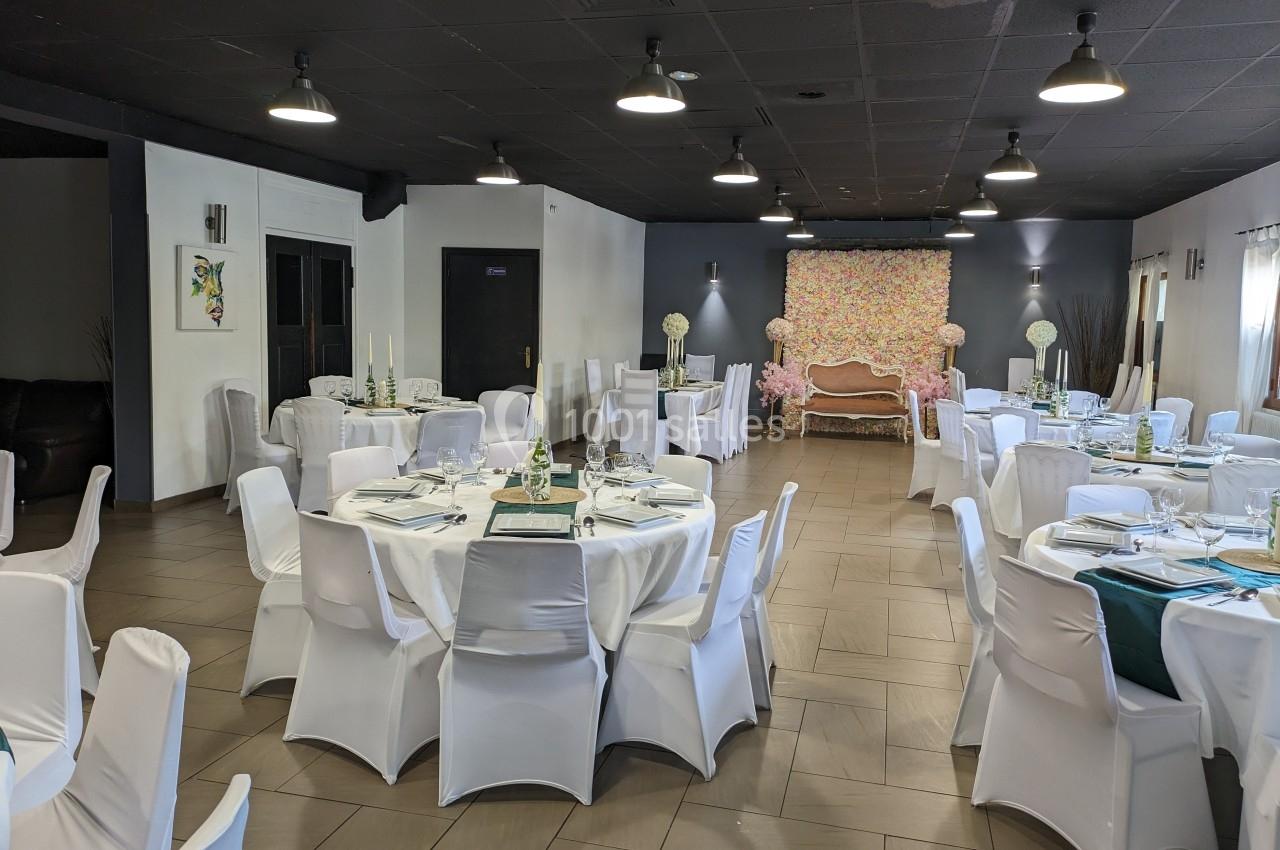 Salle de réception décorée avec des tables rondes dressées, chaises blanches et un fond floral coloré.