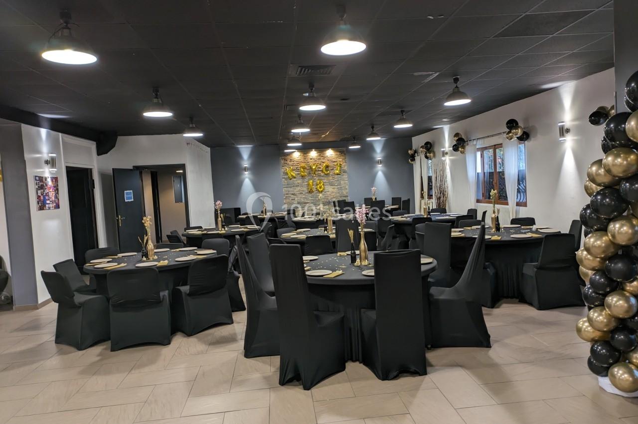 Salle décorée pour un événement avec des tables rondes noires, des ballons dorés et noirs, et un éclairage tamisé.