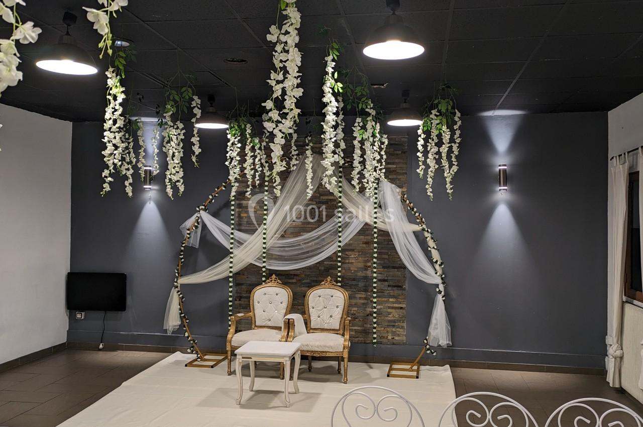 Salle décorée pour un événement avec deux chaises blanches devant une arche ornée de fleurs suspendues et de voilages.